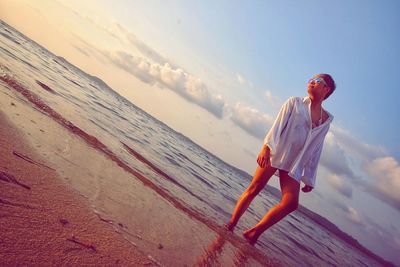 Low angle view of young woman standing at beach against sky during sunset