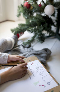 Cropped image of girl writing on paper by christmas tree at home