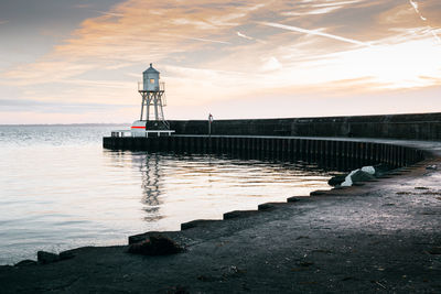 Lighthouse on pier by sea against sky during sunset