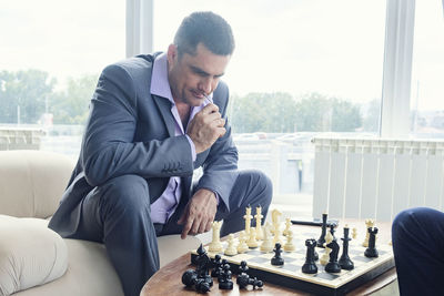 Dad and son are sitting at a table by a wide window and playing chess making moves with pieces