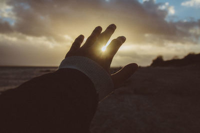 Cropped hand of person gesturing at beach against sky during sunset