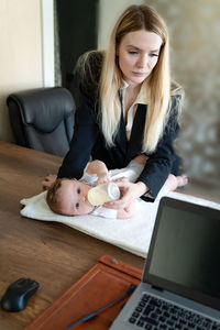 Business woman feeds baby from bottle in office, combining it with work on laptop, vertical frame