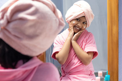 A cute indian girl child in pink bathrobe applying face cream and touching cheeks in front of mirror