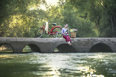Woman sitting on arch bridge over river in forest