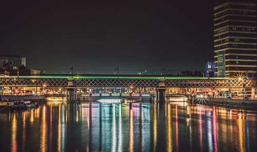 Illuminated bridge over river by buildings against sky at night