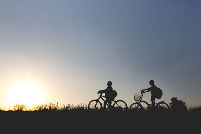 Silhouette friends with bicycles standing on grassy field against clear sky during sunset