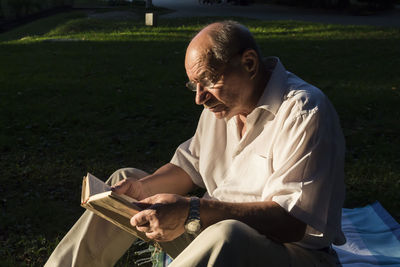 An elderly man with a watch on his hand and glasses is reading a book in a park in the fresh air.