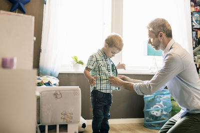 Father dressing up son in brightly lit room at home on sunny day