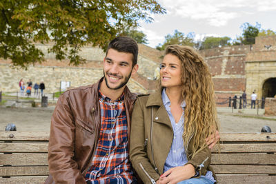 Smiling young couple sitting on bench against sky