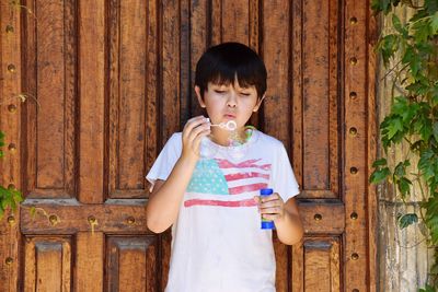 Cute boy blowing bubbles while standing against wood