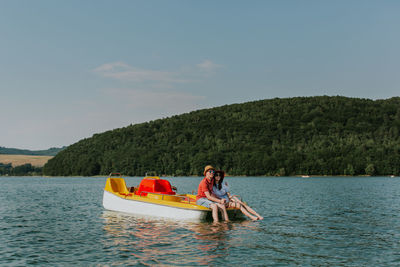 Cheerful man and woman in paddleboat on lake against sky