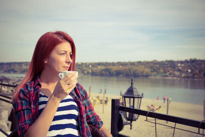 Thoughtful beautiful woman drinking coffee while standing against cloudy sky