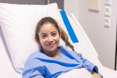 Young beautiful woman in private hospital room, dressed in blue robe, sits on bed and rejoices at