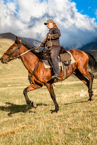 Side view of a cowgirl shepherd riding on a horse on a mountain valley against sky with clouds