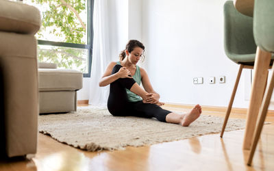 Peaceful female in sportswear practicing yoga in seated forward bend pose and stretching legs while sitting on carpet in living room