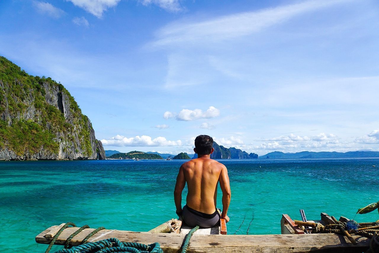 REAR VIEW OF SHIRTLESS MAN LOOKING AT SEA SHORE AGAINST SKY