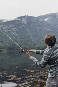 Rear view of man fishing in lake against mountains