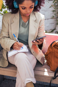 Crop young african american female manager browsing data on smartphone and writing in notebook while sitting on bench and listening to music on street