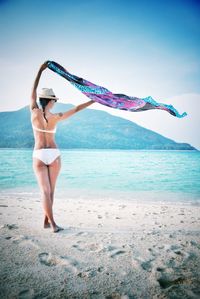 Rear view of woman standing while holding sarong aloft at beach against sky