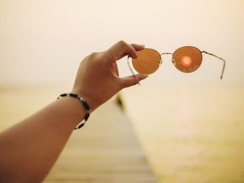 Close-up of woman hand holding sunglasses against sky at sunset