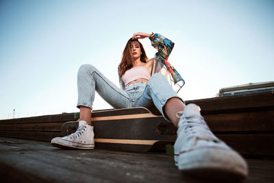 From below of stylish young women with long brown hair wearing jeans and top with sneakers sitting on wooden bench with longboard and touching head while looking at camera