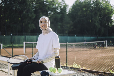 Portrait of woman wearing headscarf holding tennis racket while sitting on bench at sports court