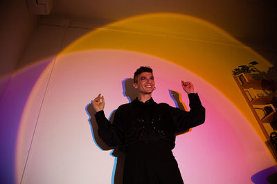 From below smiling transgender male standing arms outstretched performing in illuminated studio