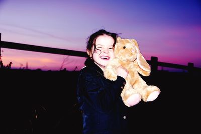 Happy girl with stuffed toy standing on field against sky at dusk