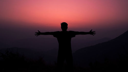 Silhouette person standing by cross against sky during sunset