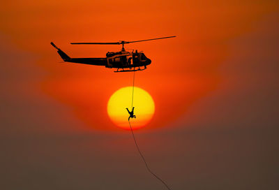 Low angle view of person hanging to helicopter against sky during sunset