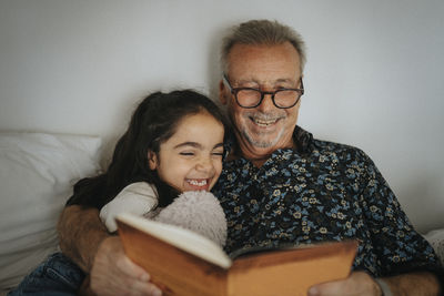 Smiling senior man reading storybook with granddaughter on bed at home