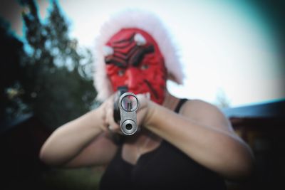 Portrait of person wearing spooky mask while holding gun against sky