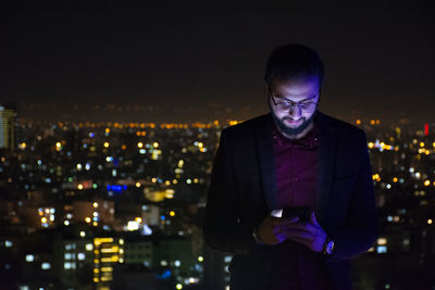 Man using mobile phone while standing against illuminated city at night