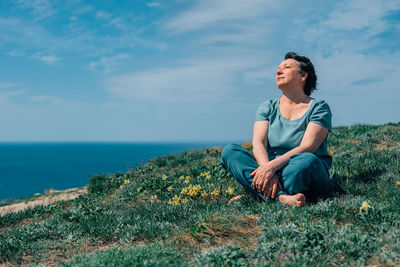 An adult woman, pensioner sits on the grass, legs crossed in a lotus position barefoot with a smile