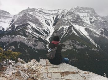 Rear view of man sitting on cliff against snowcapped mountains