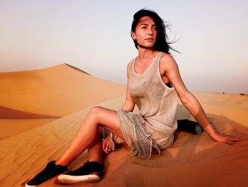 Young woman sitting on sand dune at desert against sky