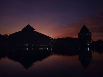 Silhouette buildings by lake against sky during sunset