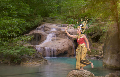 Beautiful young woman wearing crown and traditional clothing while dancing against waterfall in forest