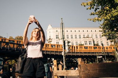 Young woman taking selfie through mobile phone while standing on street in city