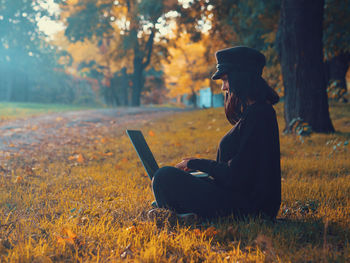 Side view of young woman using laptop while sitting at public park during autumn