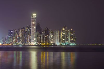 Illuminated modern buildings by river against sky at night