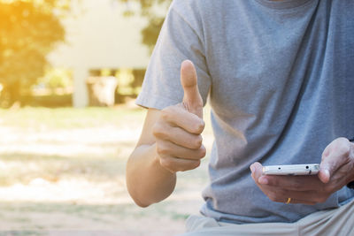 Midsection of man gesturing thumbs up while holding mobile phone outdoors