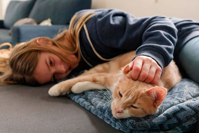 A young adolescent girl lying on a couch finds comfort by snuggling and petting her tabby cat