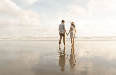 Rear view of couple holding hands at beach against sky
