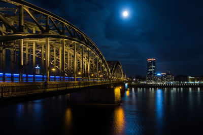 Illuminated bridge over river by buildings against sky at night