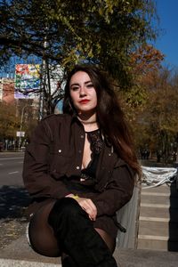 Portrait of young woman sitting against trees in city