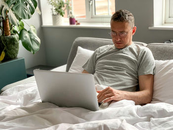 Middle aged man using laptop at home in bed vibes white bedsheets early morning working freelancer