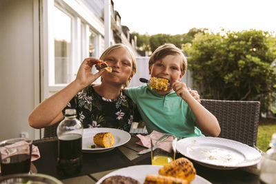 Portrait of siblings eating grilled corn and meat while sitting in back yard