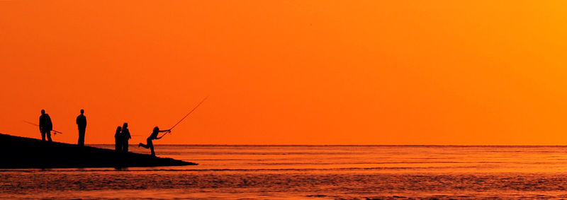 Silhouette people fishing on beach against orange sky