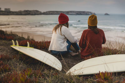 Female friends sitting with surfboards at beach during sunset
